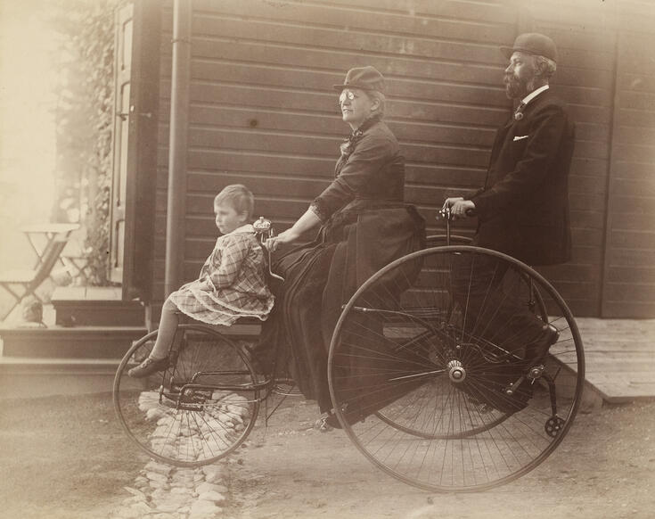 En ovanlig cykel med ett litet framhjul och ett stort bakhjul. På cykeln åker en liten flicka längst framme, i mitten flickans mor i en lång kjol och längst bak flickans far.