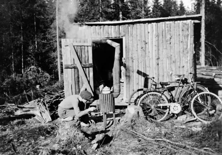 En bod av bräder med öppen dörr. Framför boden sitter en man på huk och håller på att tända i en flyttbar kamin.  Några cyklar lutar mot boden. 