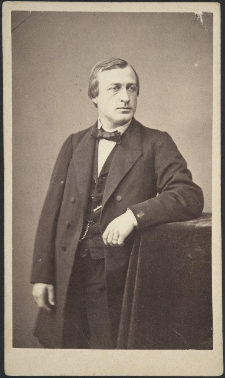 En man iklädd en knälång rock, väst och rosett står bredvid ett högt bord täckt med en sammetduk och stöder den vänstra armen på bordet. På västen har fästs en fickurskedja.