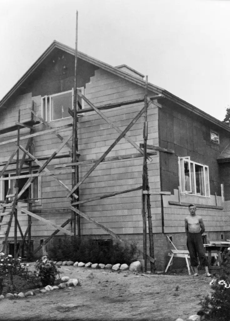 Vid frontmannahusets gavel finns en byggnadsställning. Husets fasadbeklädnad håller på att monteras. Bredvid huset står en man med bar överkropp.