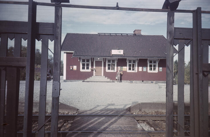 En rödmålad stationsbyggnad i en våning, med fyra fönster med sex rutor i fasaden. Byggnadens hörn och karmar är vitmålade, medan ytterdörren är brun. På väggen till huset finns en brevlåda, där en pojke eller man lämnar en försändelse.