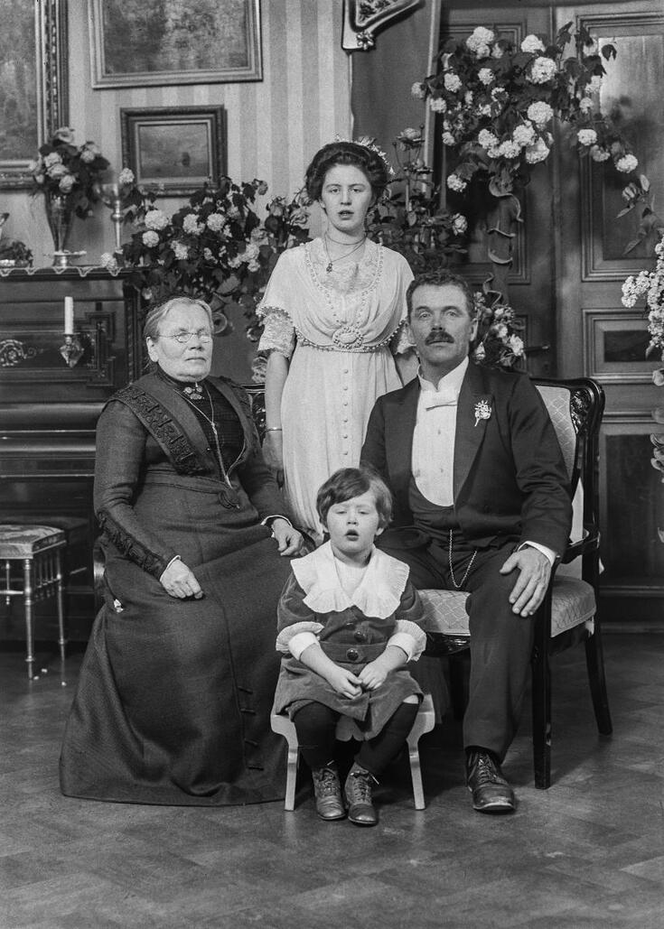 Vasemmalla istuu tummiin pukeutunut vanhempi nainen ja hänen vieressään mies. Heidän takanaan seisoo vaaleaan pukeutunut nainen ja edessään istuu pienellä tuolilla tyttö. Taustalla näkyy kukkakoristeita, piano ja seinällä olevia tauluja.