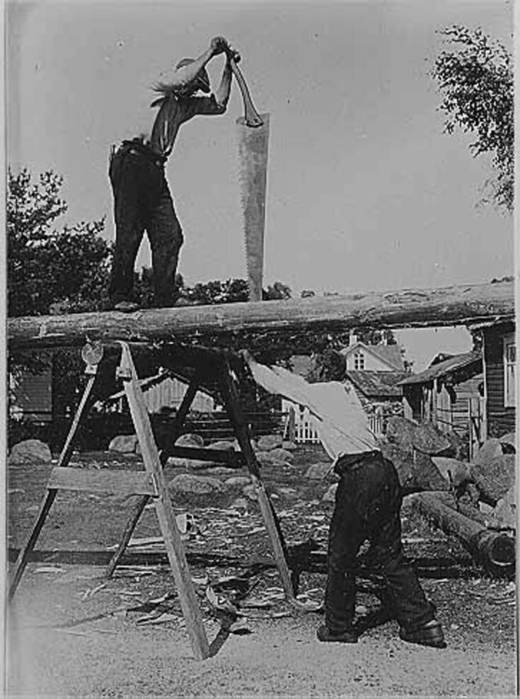 Kaksi miestä työstää tukkia laivan laudoitukseksi. Toinen seisoo pukin varaan nostetulla tukilla ison työkalun kanssa, toinen seisoo maassa ja tukee käsillään tukkia.