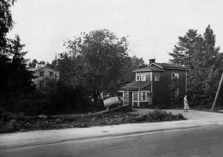 Kuvassa oikealla ja vasemmalla puurakennukset, niiden välissä omenapuu. Etualalla tynnyri kärryjen päällä, etuoikealla nainen katsoo olkansa yli kuvaajaa. Tie kulkee etualalla.