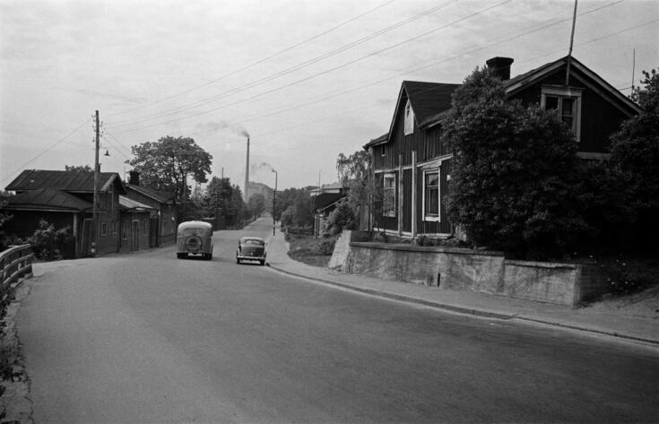 Hämeentietä reunustavat matalat puurakennukset. Tiellä näkyy yksi auto ja linja-auto. Taustalla tehtaat savuavine piippuineen.