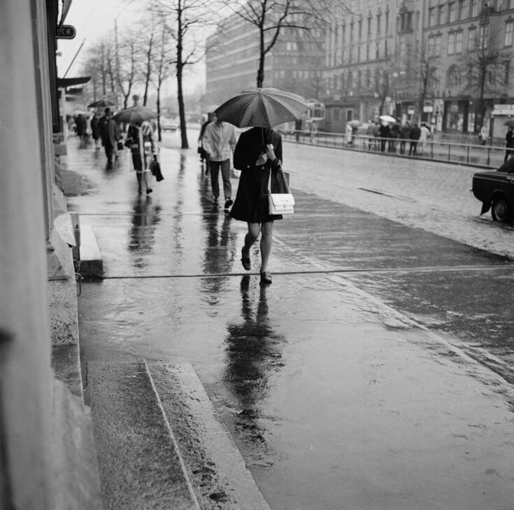 Människor promenerar på en regnig trottoar. I bakgrunden Mannerheimvägen 5, 7, 9.
