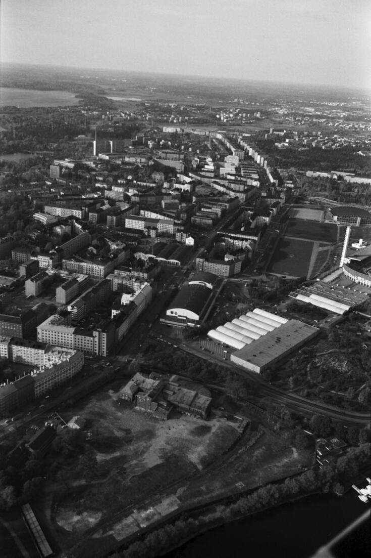 Mannerheimintie. Viistoilmakuva pienlentokoneesta Töölönlahden suunnalta koilliseen. Etualalla Töölön sokeritehtaan tontti, jonka Helsinginkadun laidalla tehdasrakennus. Keskellä Messuhalli, oikealla Mäntymäenkentällä väliaikaiseksi rakennettu  C-Messuhalli eli Kansojen halli. Taustalla keskellä Mannerheimintietä.