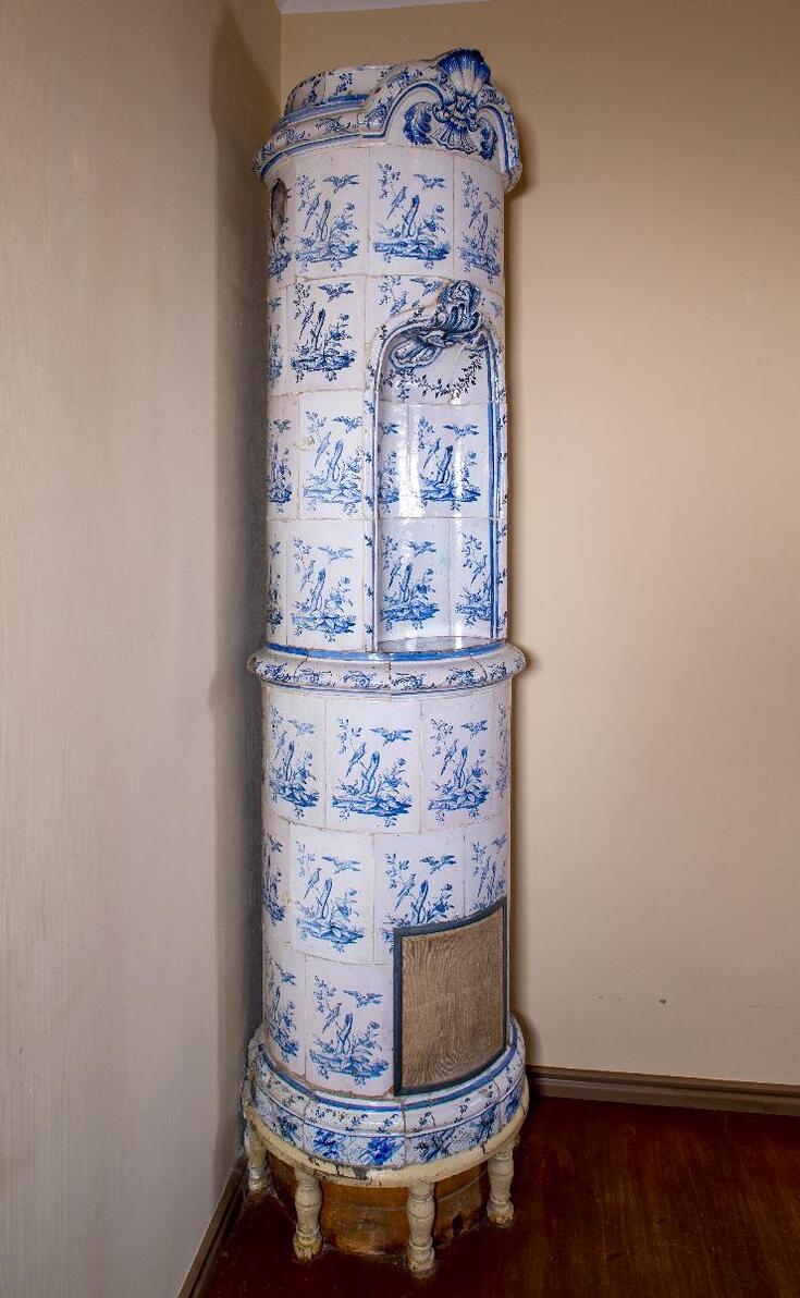 Lieriömäinen uuni, jonka kaakelit on koristeltu puunrungolla, kukilla ja linnuilla. Sininen kiinalaisvaikutteinen maalaus lasitteen alla. Sorvatuilla jaloilla, kaksiosainen, ylemmässä pyöreä syvennys. Syvennyksen pohja koostuu erikuvioisesta kaakelista: viljalyhde, harava ja viikate.