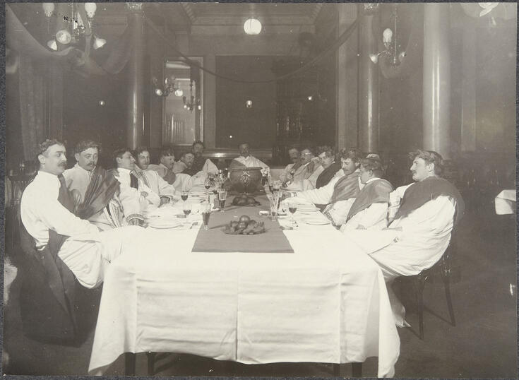 Ryhmä miehiä istuu pitkän valkoisella liinalla peitetyn pöydän ääressä. Jokaisella on laakeriseppele päässä ja yllään tooga. 