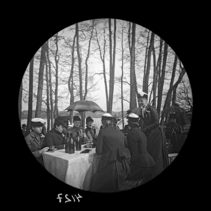 Puistoon on katettu pöytä, jonka päällä on pöytäliina ja pulloja. Pöydän ääressä on miehiä ja naisia. Joillakin naisilla on ylioppilashattu, jollaisia käytettiin vain 1890-luvulla.