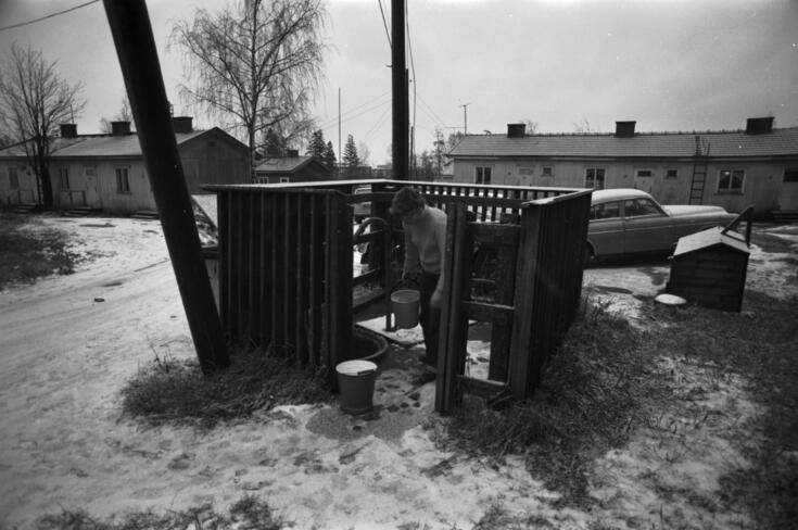 Pienen aitauksen ympäröimä vesiposti, jonka ääressä on nainen kahden ämpärin kanssa. Maassa on lunta. Taustalla näkyy matalia parakkitaloja ja vuosikymmeniä vanha auto.