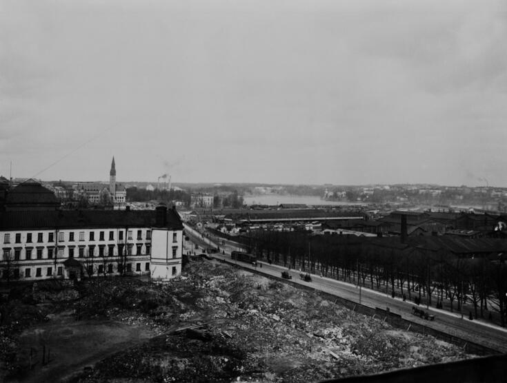 I förgrunden syns en tom tomt med ruiner av en kasern. I bakgrunden till vänster Nationalmuseet. I mitten löper nuvarande Mannerheimvägen. Till höger Tölö godsstation och gasverket.