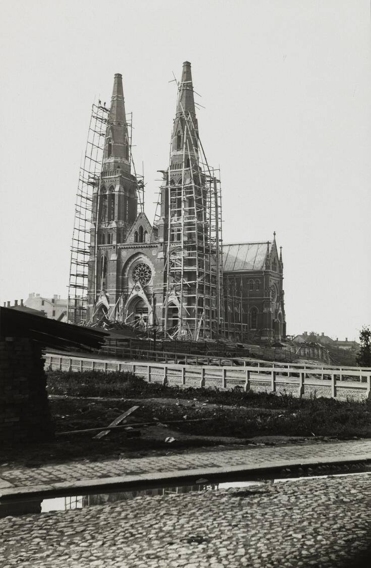 Etualalla mukulakivikatua ja sadevettä, taustalla kohoaa rakennustelineiden ympäröimä Johanneksen kirkko, jonka rakentaminen on loppusuoralla. Kirkkoa ympäröivät puiset rakennustelineet.