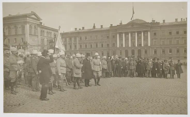 Iso joukko seisoo Senaatintorilla löyhässä rivimuodostelmassa. Suurin osa paikallaolijoista on pukeutunut sotilasuniformuihin ja useammalla on valkoiset karvalakit päässä. Ihmisjoukossa näkyy myös muutamia silinteripäisiä siviilejä, joista muutamilla on valkoiset käsivarsinauhat. Joukon takarivistä nousee valkoinen lippu. Taustalla näkyy valtioneuvoston linna.