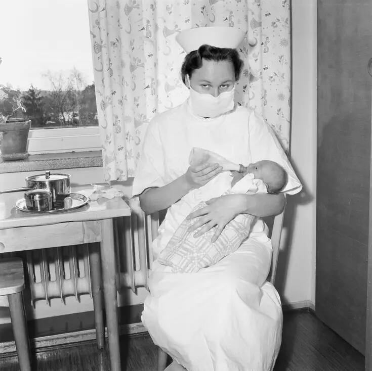 En sköterska klädd i arbetsuniform sitter vid ett bord som står framför ett fönster och matar en baby med nappflaska. Sköterskan tittar mot kameran, och hon bär mun-nässkydd.