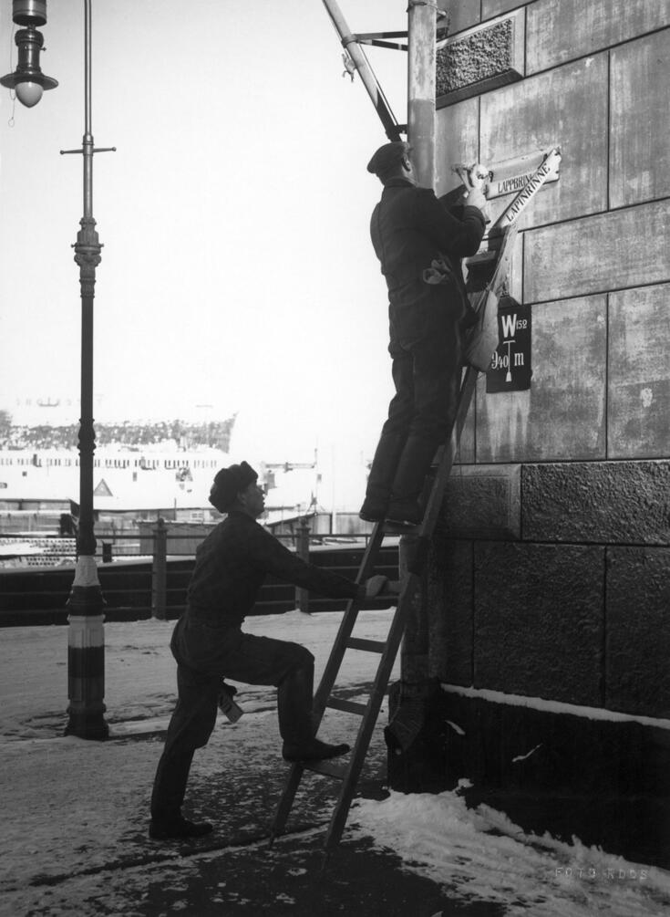 Mies seisoo tikapuilla kiinnittämässä kadunnimikilpeä talon seinään. Toinen tukee tikkaita.