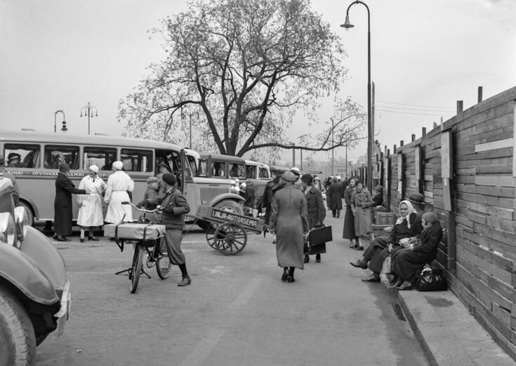 Bussar och passagerare på Salomonsgatan och nuvarande Paasikiviplatsen. Bakom planket till höger har man rivit den norra flygeln av Åbo kasern som förstördes i inbördeskriget 1918. Den ersattes av Glaspalatsets biograf Rex år 1936. Trädet som visas på bilden är en så kallad knäckepil som skyddades år 1924. Det föll omkull i en storm 29.12.2003. Det ersattes av ett träd som drivits upp från en stickling av det fallna trädet.