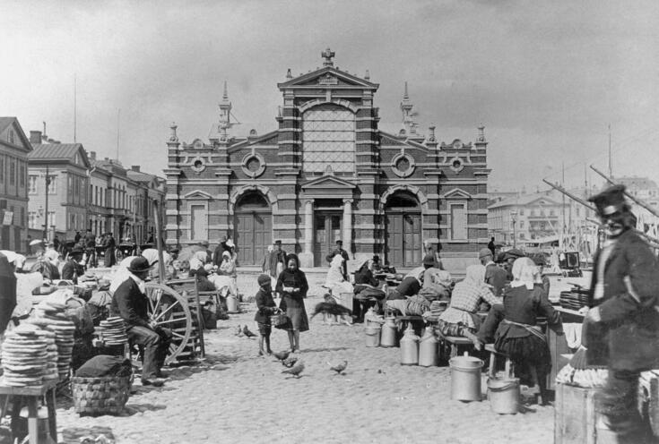 : På torget utanför ingången till saluhallen finns försäljare med sina produkter. I mitten ser man några duvor och en liten pojke och en flicka med en korg. Barnen kanske matar duvorna.