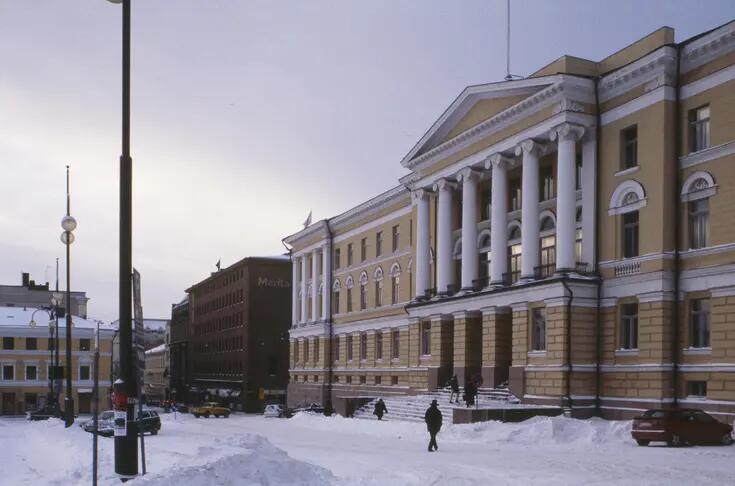 Etualalla näkyy keltaisen hallinnollisen rakennuksen julkisivu. Vuodenaika on talvi ja kadulla on lunta. Muutama henkilö käyskentelee rakennuksen portaiden tuntumassa.