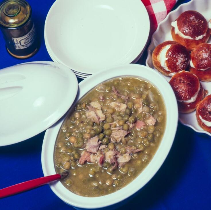 Keittokulhollinen hernekeittoa, jossa on paljon sianlihaa. Vieressä pn pino valkoisia lautasia sekä vadillinen laskiaispullia.