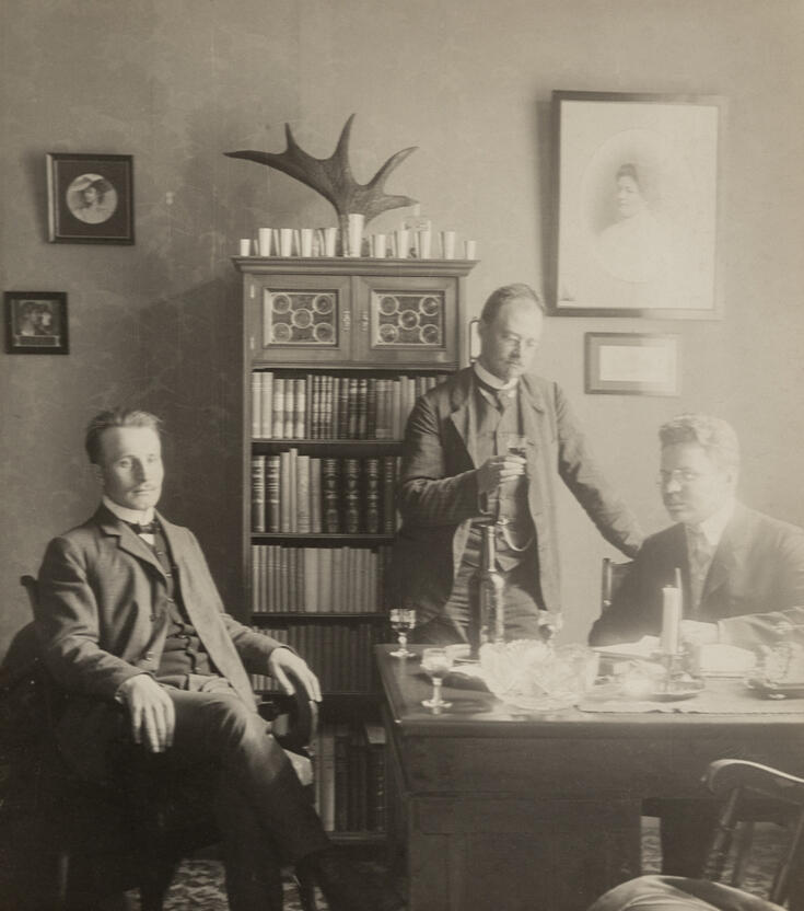 Tre män dricker vin i ett arbetsrum. Till vänster sitter en man på en stol, i mitten syns en bokhylla med pokaler och ett älghorn. Framför hyllan står en man med ett glas vin i handen. Till höger om honom sitter en man med glasögon bakom ett skrivbord. På bordet finns brevpapper, en ljusstake med ett ljus, en kristallskål, en vinflaska och tre glas. I förgrunden står en tom stol, på vilken fotografen och ägaren av arbetsrummet sitter.