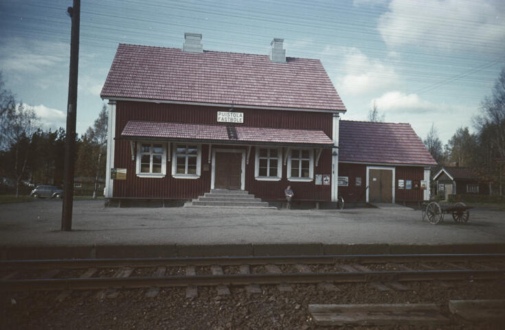 Punainen, puusta rakennettu harjakattoinen 1,5-kerroksinen asemarakennus, jonka seinustalla on penkki, jolla istuu mies.