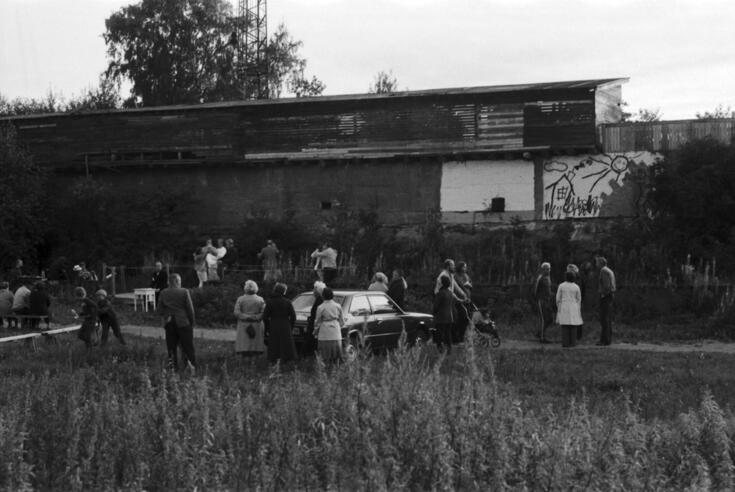 Hiidenkivenpuisto. Ihmisiä tanssimassa "uudella" Mosabackan lavalla sekä seisoskelemassa horsmia kasvavalla maalla (Hiidenkivenpuisto). Taustalla urheilutalon lähistöllä olevaan todennäköisesti varastoalueeseen liittyviä rakennelmia. Alkuperäinen  Mosabackan lava sijaitsi Sanduddin tapettitehtaan naapurissa, Viertolantien varrella.