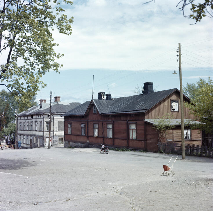 Kesällä otetussa valokuvassa on vasemmalla vaalea puutalo ja oikealla on ruskea puutalo, jonka edessä on mopo. Kuvassa etualalla on nukenvaunut.