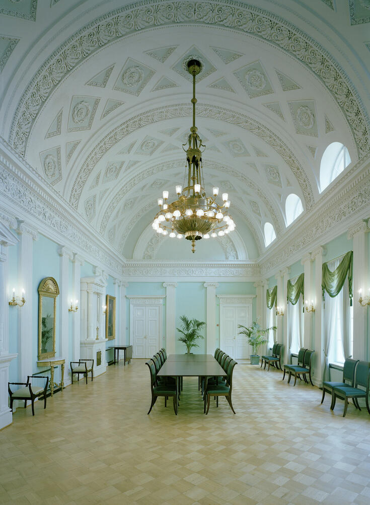 En öppen sal med ett vitt, högt och dekorativt valvtak, ljusturkosblåa väggar, parkettgolv med diagonalt rutmönster, gyllene vägglampetter och en stor gyllene lampkrona samt väggspeglar med guldramar. I mitten av salen finns ett långt bord och vid dess långa sidor stolar.