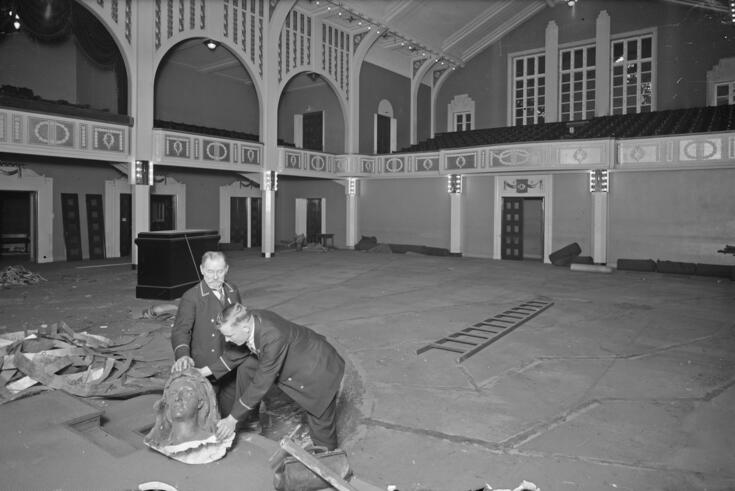 Tyhjä sali, jonka lattialla makaa tikapuut. Etualalla kaksi vahtimestaria on pakkaamassa veistoksen päätä.