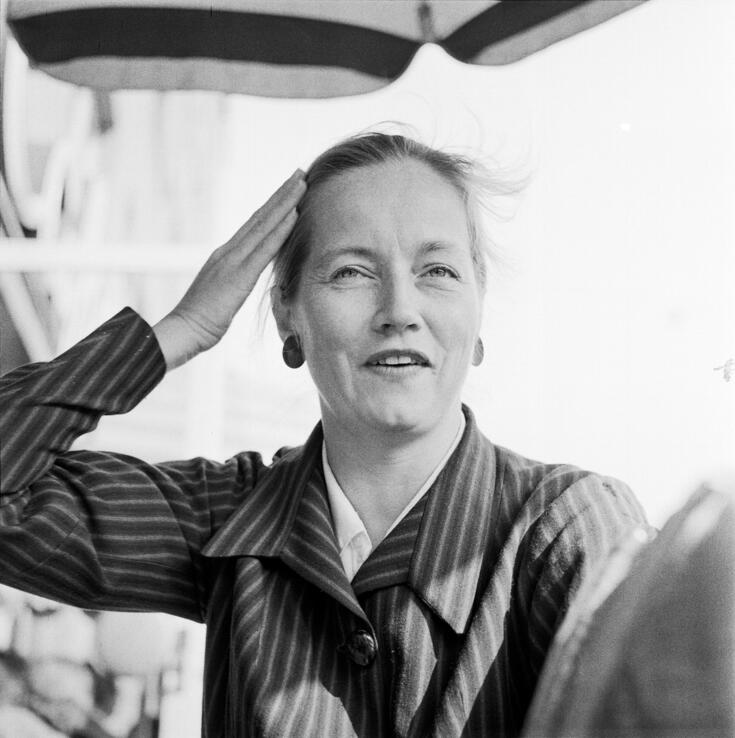 En medelålders kvinna ler mot kameran. Hon håller sin högra hand bakom sin högra tinning. Hon är klädd i en randig blus eller jacka.