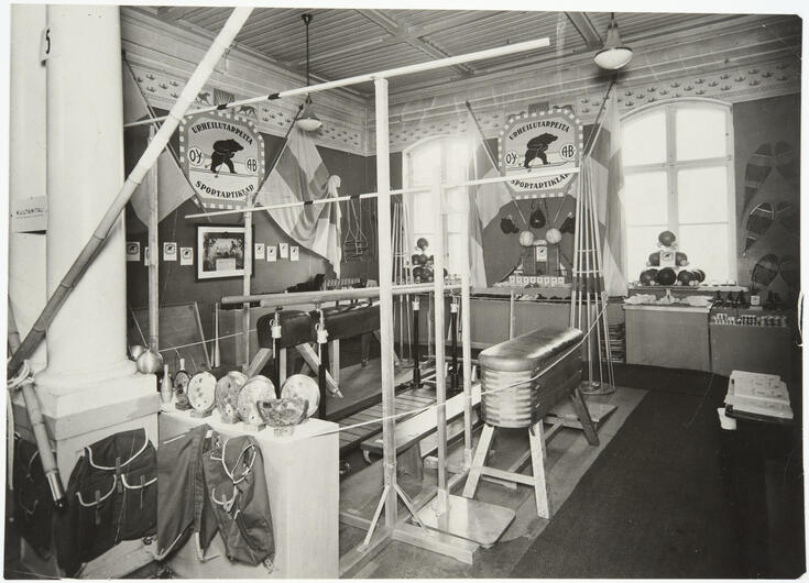 Idrottsartiklar har ställts ut i en skolbyggnad. På väggarna hänger Finlands flaggor och firmaskyltar med en tecknad skidande björn och texten O.Y. Urheilutarpeita - A.B. Sportartiklar.