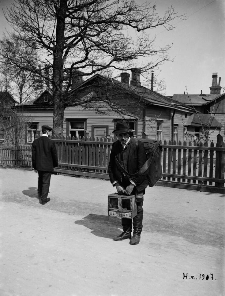Posetiivari seisoo lumisella tiellä posetiivi käsissään ja reppu selässään. Hänen takanaan on lauta-aita ja puutaloja. Hänen vasemmalla puolellaan seisoo mies selin kuvaajaan.