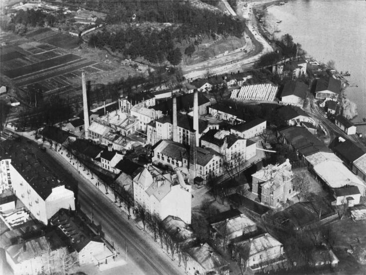 Flygbilden visar ett fabriksområde på stranden till Tölöviken. På norra sidan av området syns åkrar.