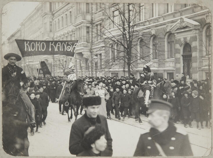 Polyteekkarien karnevaalikulkue kävelee ja ratsastaa keskellä Bulevardia. Ympärille on kokoontunut runsaasti pikkupoikia katsomaan.
