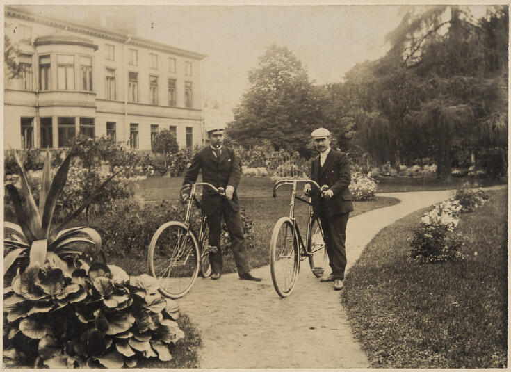 Två unga med med varsin cykel på stråket i parken. Bakom dem syns en byggnad på två våningar. Runt dem syns växtlighet.