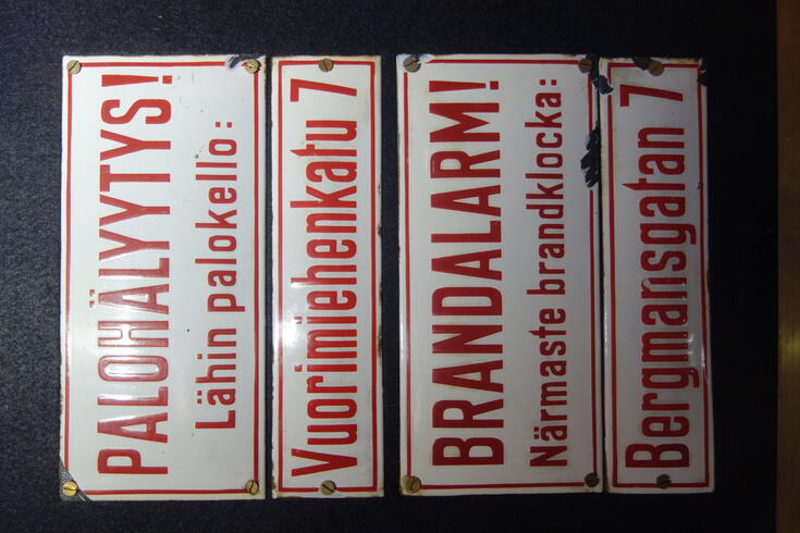På en vägg fästa emaljskyltar med röd text på vit botten: Palohälyytys! Lähin palokello: Vuorimiehenkatu 7. Brandalarm! Närmaste brandklocka: Bergmansgatan 7.
