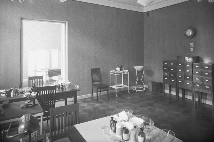 Mottagningsrum på ett sjukhus. I förgrunden syns ett bord med glasflaskor och annan utrustning, till vänster ett sidobord med mikroskop. Bakom det står ett skrivbord med stol för läkaren och patienten, till höger ett ställ med tvättfat samt ett dokumentskåp