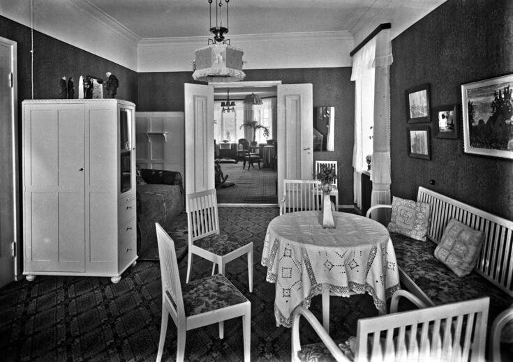 Ett rum med möbler såsom bord, stolar, soffa och skåp. Genom en öppen pardörr syns ett annat rum som hör till bostaden.