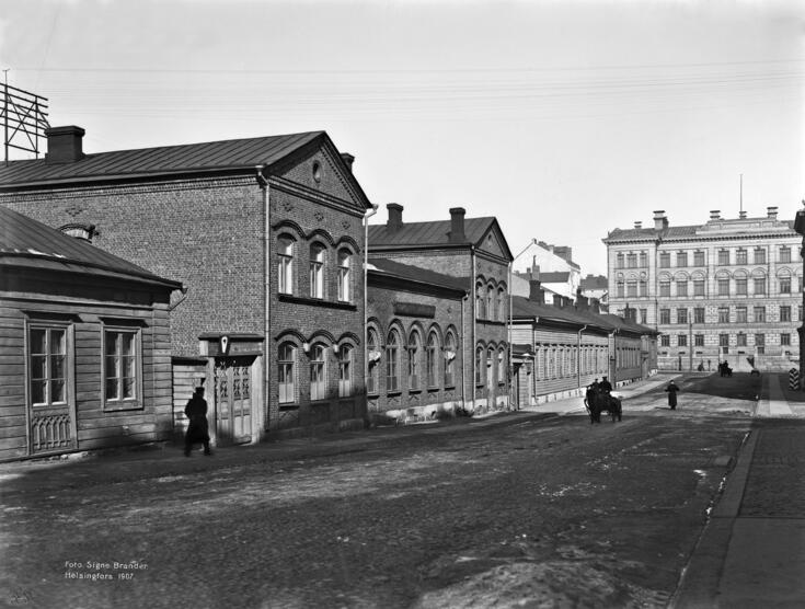 Etualalla kulkee tie. Sen varrella on matala puutalo, kaksikerroksinen tiilitalo, ja matala puutalo. Talojen edessä kävelee muutama ihminen. 