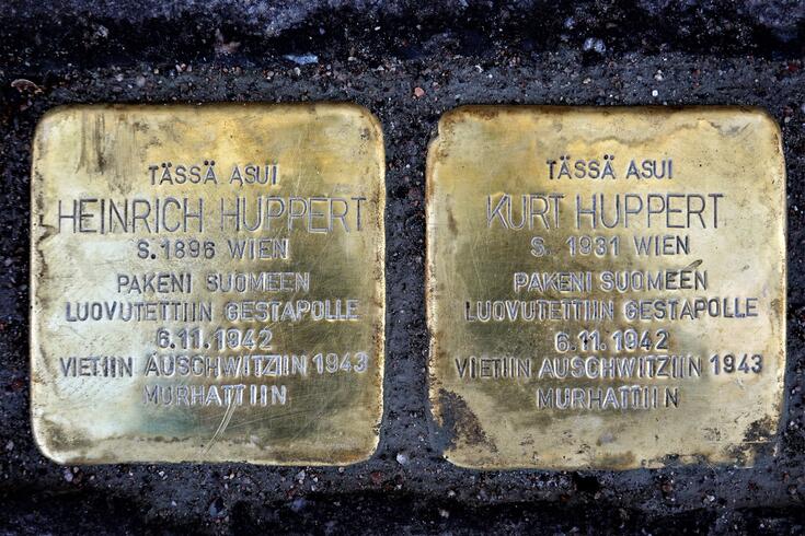 Kaksi metallilaattaa asfaltissa. Niissä tekstit: Tässä asui Heinrich Huppert s 1896 Wien Pakeni Suomeen Luovutettiin Gestapolle 6.11.1942 Vietiin Auschwitziin 1943 Murhattiin