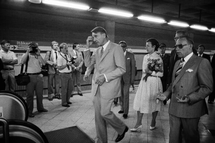 Presidentti Koivisto kävelee Itäkeskuksen metroaseman laiturilla Metrojohtaja Unto Valtasen vierellä. Taustalla presidentin puoliso Tellervo Koivisto kantaa kukkakimppua. Joukko toimittajia ottaa valokuvia.