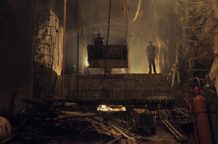 Kaksi työmiestä seisoo hämärässä, vielä rakenteilla olevassa metrotunnelissa. Toinen heistä seisoo maansiirtokoneen kauhassa.