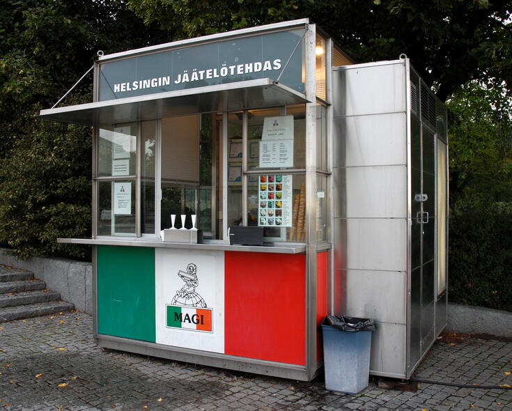Försäljningsståndet är av glas och metall, ovanför takskägget finns texten Helsingin jäätelötehdas. Den främre väggen nedanför försäljningsdisken har tejpats med den italienska flaggans färger.