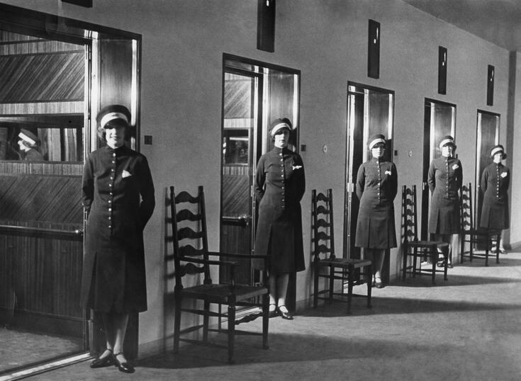 Fem uniformsklädda kvinnor står vid fem öppna hissdörrar. Mellan hissarna finns stolar.