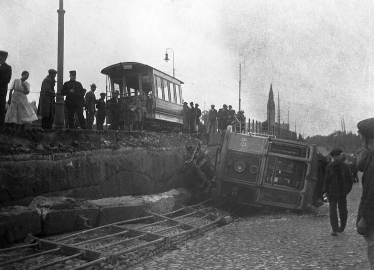Raitiovaunun etuvaunu kaatuneena. Toinen vaunu on pystyssä kiskoilla, ihmisiä katsomassa kaatunutta vaunua.