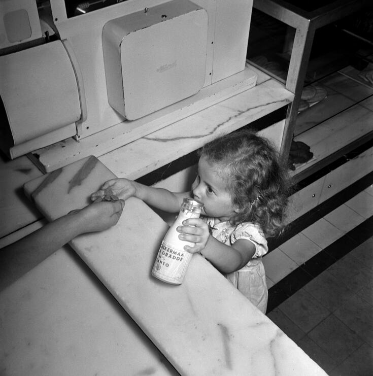 Pieni tyttö kermapullo toisessa ja kolikoita toisessa kädessä tiskin ääressä ylhäältä kuvattuna. Myyjän käsi ottamassa vastaan kolikoita.