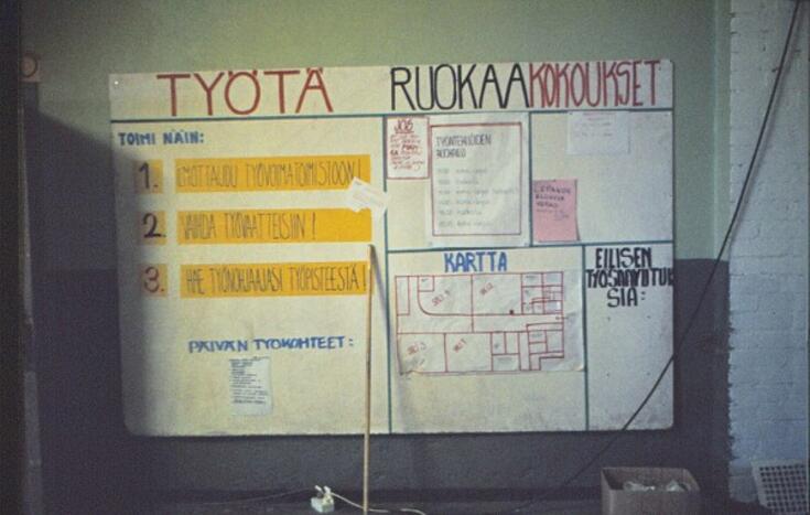 Anslagstavla i det ockuperade Lepakkoluola med anvisningar för deltagande i talkot. På tavlan står rubrikerna "TYÖTÄ", "RUOKAA" och "KOKOUKSET", alltså ARBETE, MAT och MÖTEN. Listor med dagens arbetsuppgifter, föregående dags färdiga arbeten och en karta över byggnaden.
