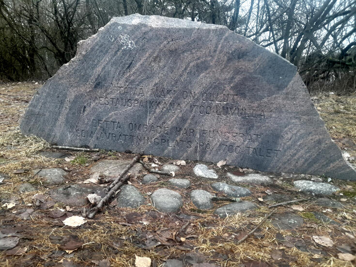 Kivipaasi, jossa teksti TÄMÄ MÄKI ON OLLUT MESTAUSPAIKKANA 1700-LUVULLA DETTA OMRÅDE HAR FUNGERAT SOM AVRÄTTNINGSPLATS PÅ 1700-TALET.