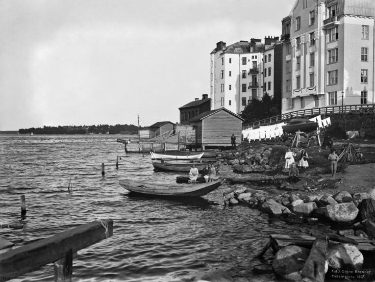 Kuvassa näkyy merenranta, rantakallioita ja oikealla nousevat kivitalot. Rannalla on kaksi puista rakennusta ja niiden edustalla vedellä puuaidoilla rajatut alueet. Rannalla on pyykkejä kuivumassa.